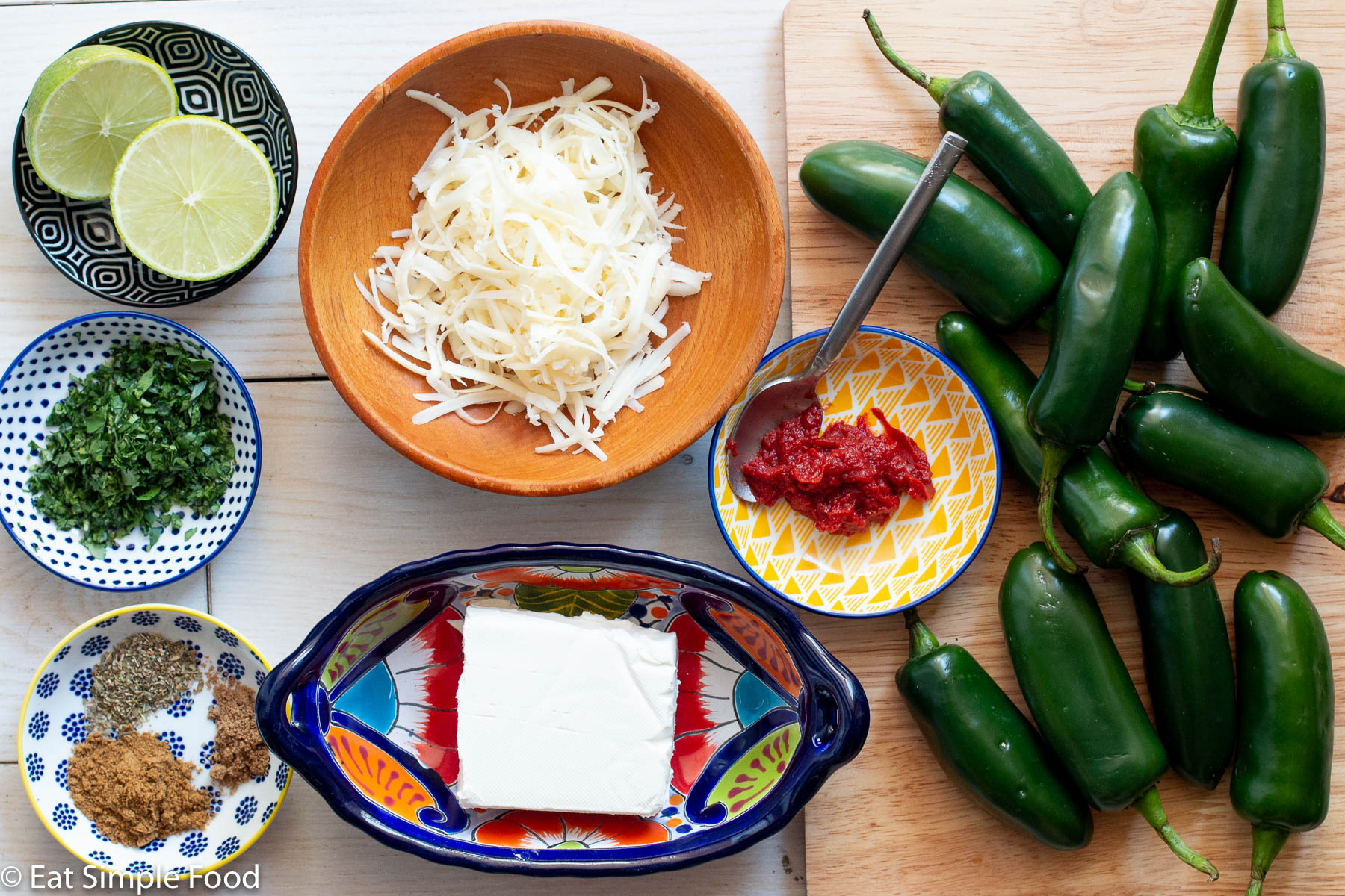 Top view of ingredients: containers each of cream cheese block, spices, tomato paste with spoon, grated white cheese, chopped cilantro, 2 lime halves, and 12 whole jalapenos.