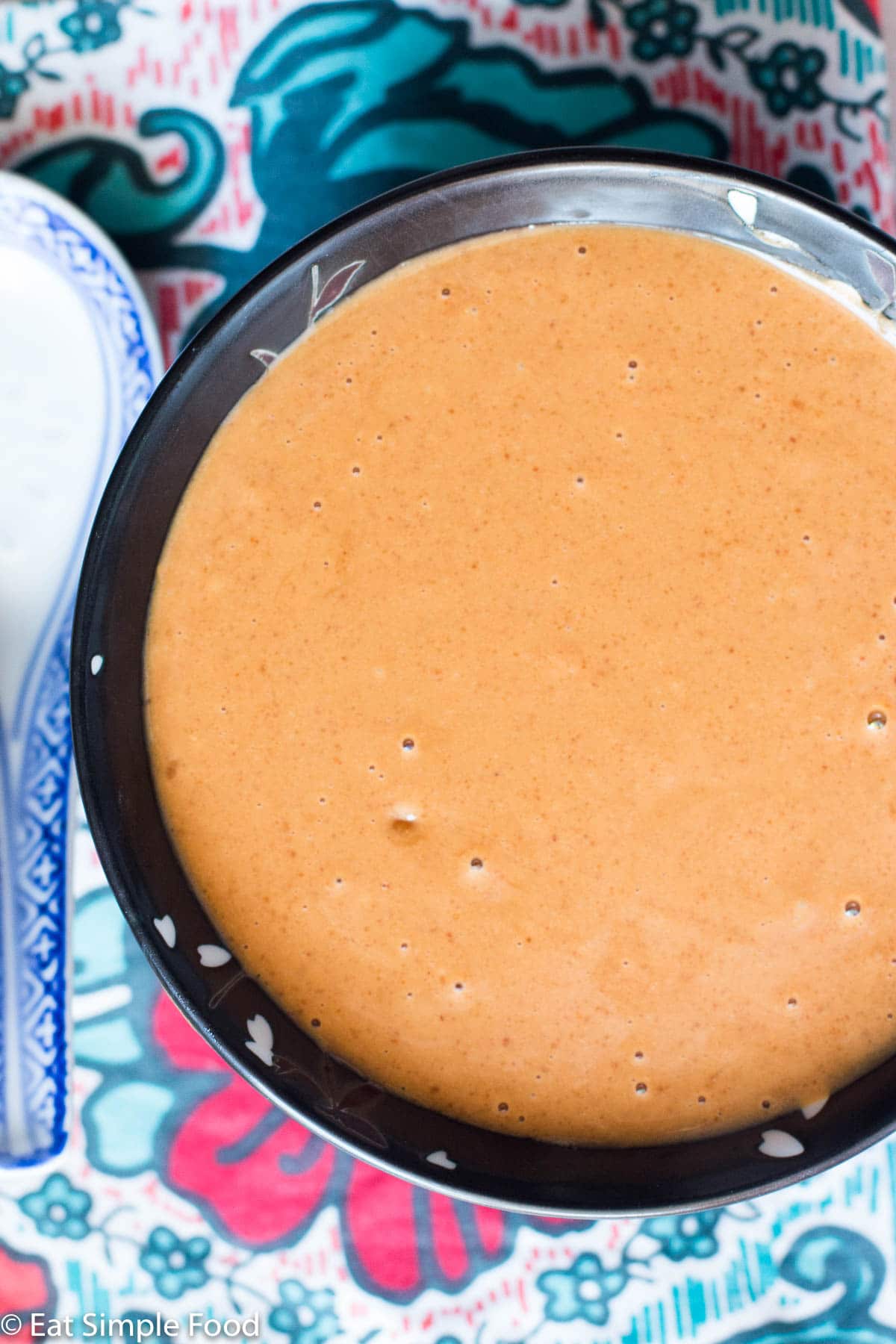 Black Bowl of Brown Peanut Sauce with a white chinese style spoon in it. On a colorful placemat. Top view.