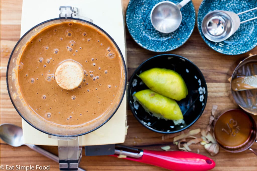 Food Processor full of brown sauce with discarded ingredients of two ½'s of a lime, ginger peel, and peanut butter in a bowl.