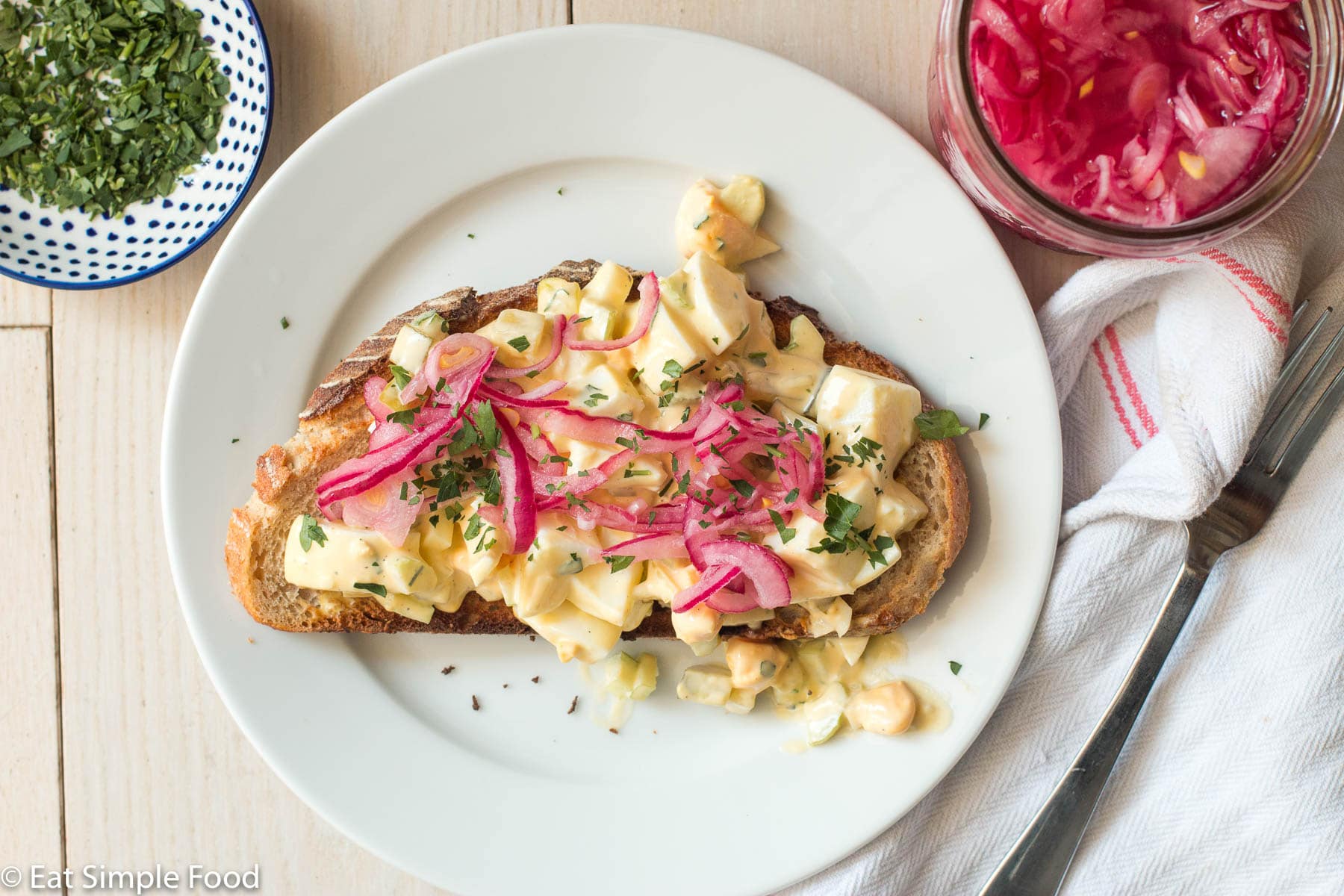 Top view of toast topped with a chunky egg salad and pickled red onions on a white plate with napkin and fork on the side.
