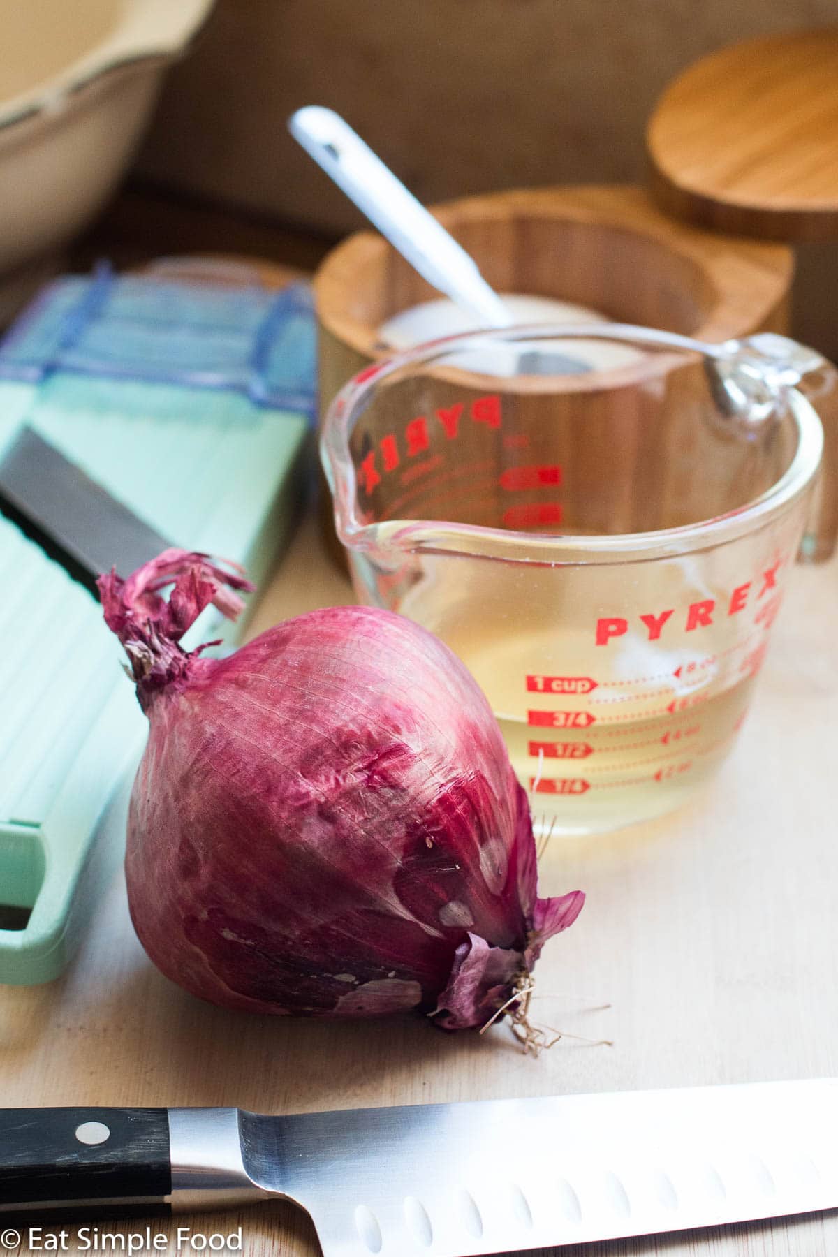 Ingredients on a wood cutting board: whole red onion, measuring cup of ¾ cider vinegar, green mandolin, chef's knife, and wood bowl of salt.