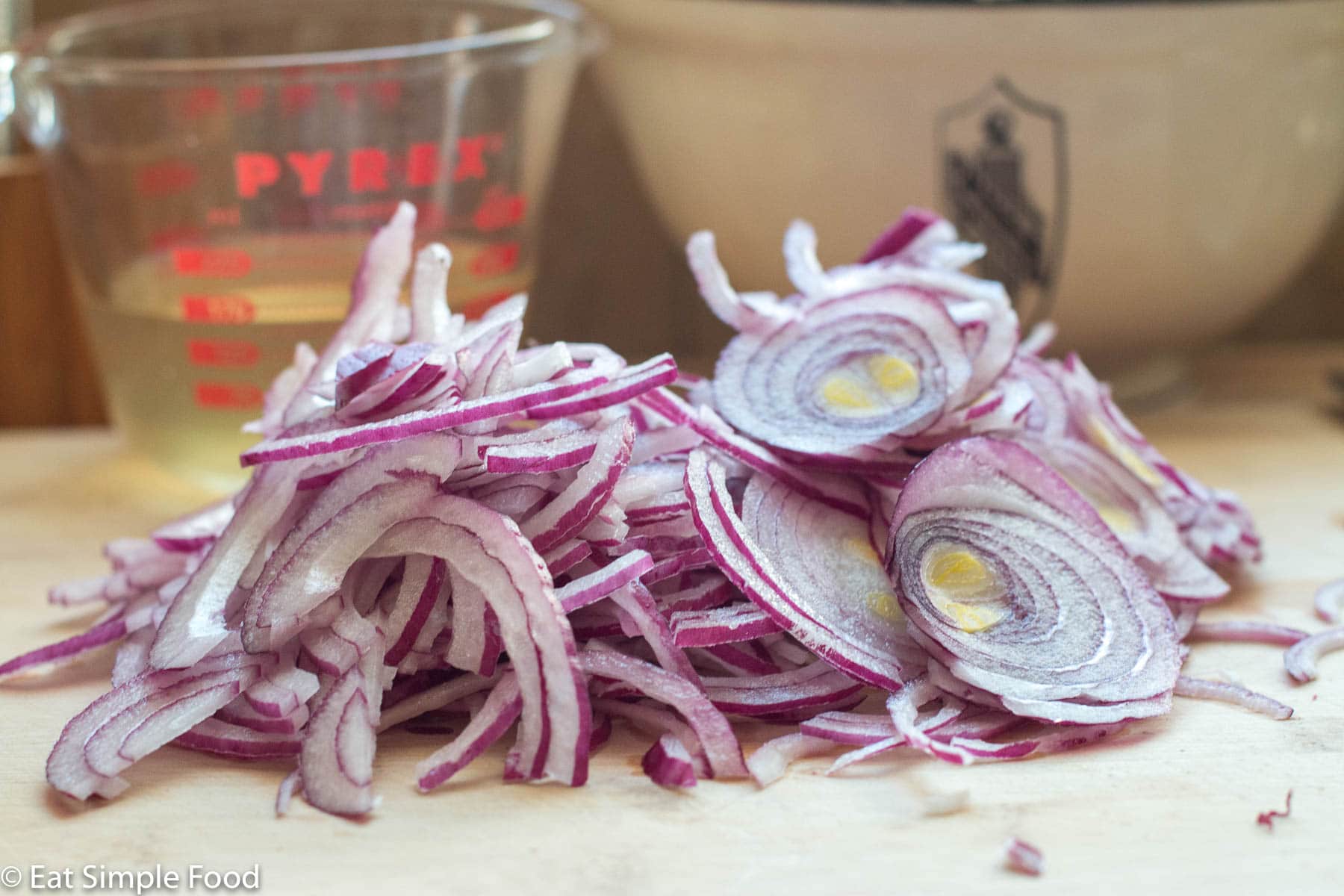 A pile of thin sliced red onions on a wood cutting board with a measuring cup of vinegar in the background.