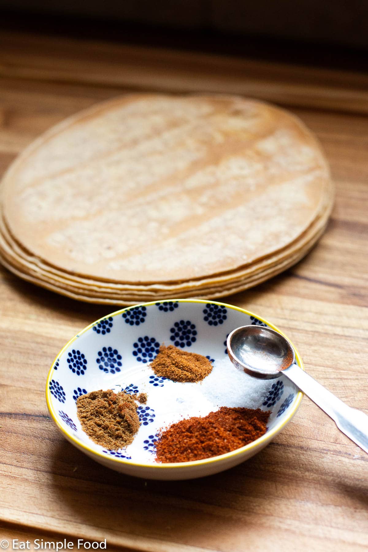 Raw uncut tortillas on a wood cutting board with a small bowl of red, brown, and yellow spices in the front.