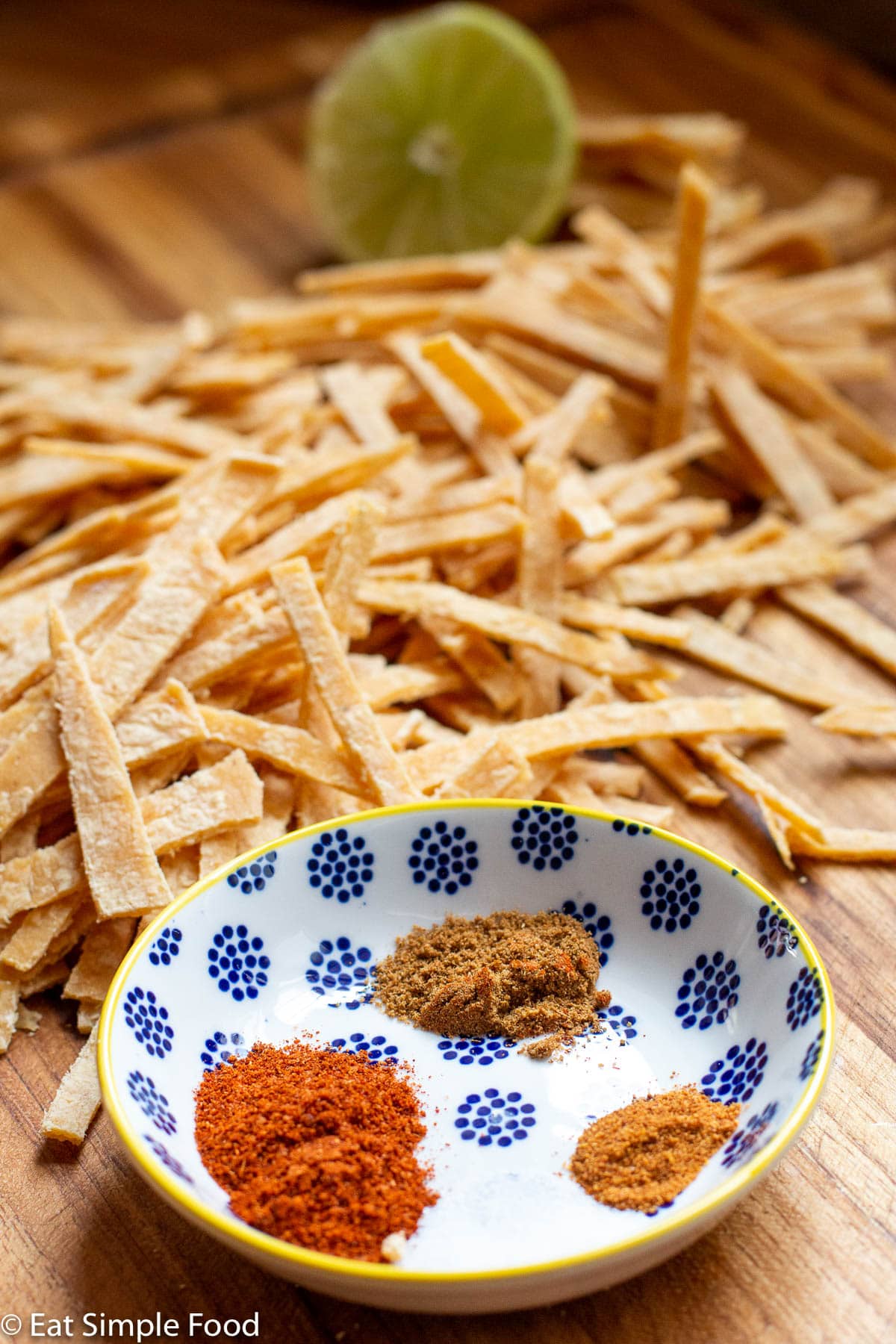 Raw tortilla strips on a wood cutting board with ½ a lime in background and a small bowl of red, brown, and yellow spices in the front.