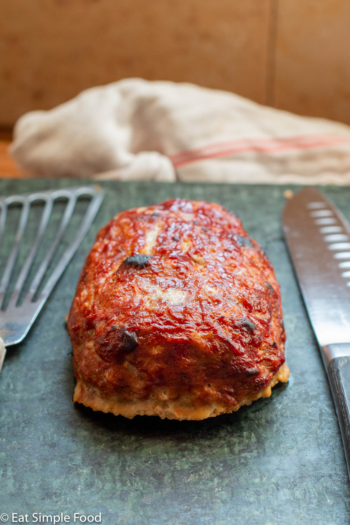 Side view of turkey meatloaf with a brown red ketchup glaze. Knife and metal spatula on the side. White towel in the background.