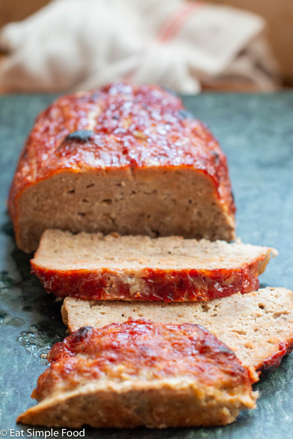 Side view of sliced turkey meatloaf with a brown red ketchup glaze on a dark green cutting stone.