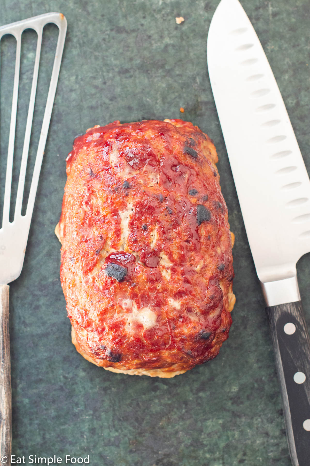 Top down view of turkey meatloaf with a brown red ketchup glaze. Knife and metal spatula on the side.