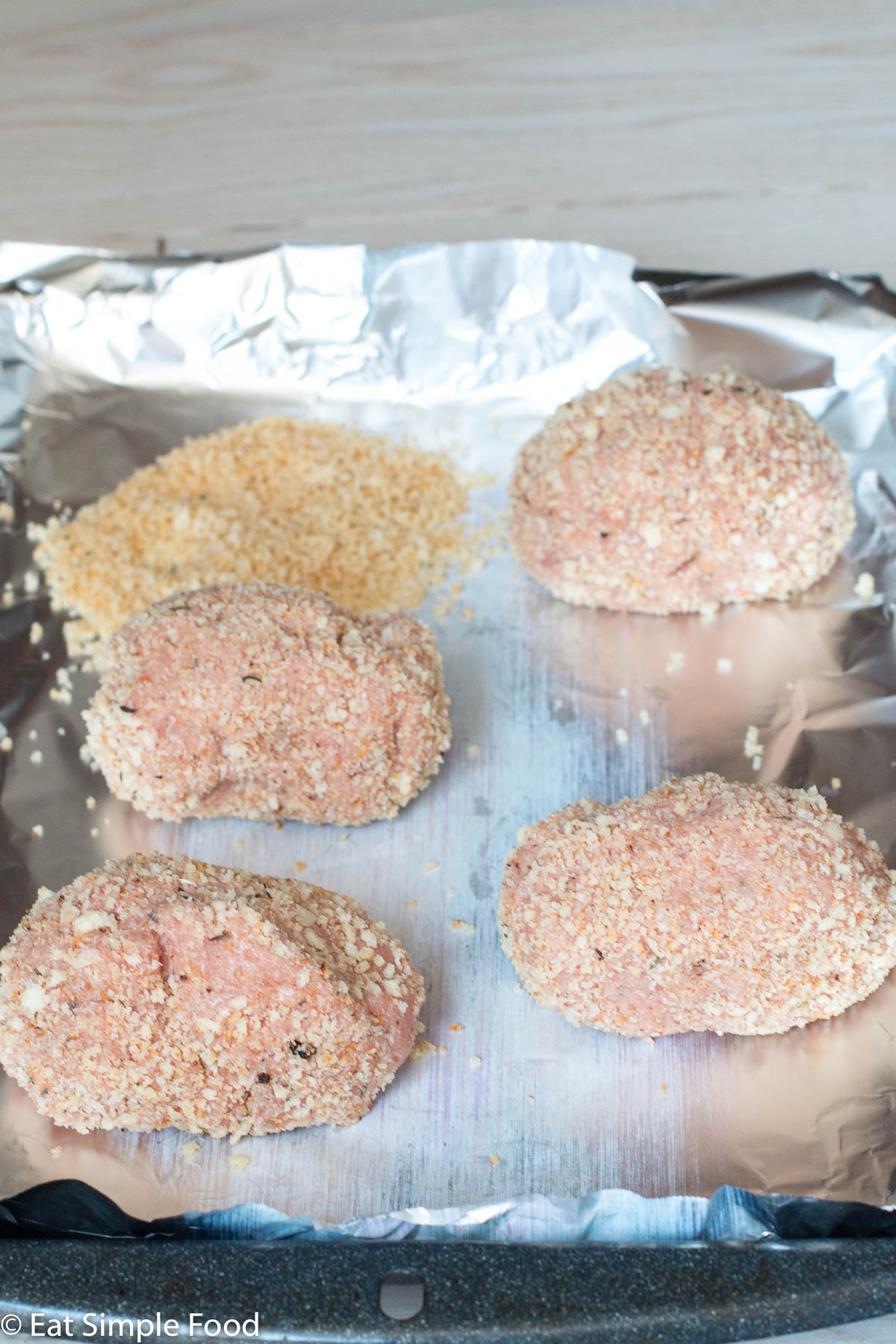 4 uncooked raw turkey scotch eggs on an aluminum lined baking sheet with breadcrumbs on it as well.