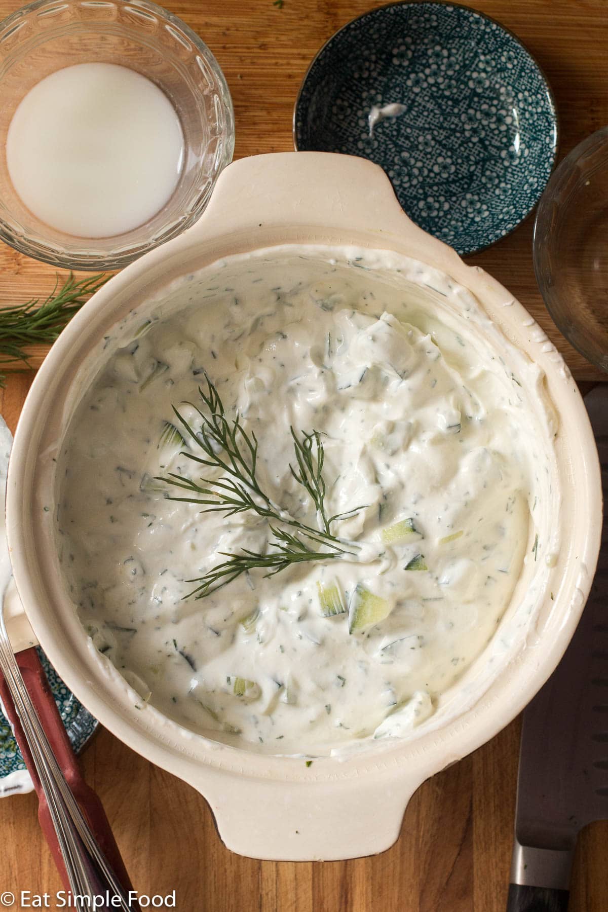 Top view of white bowl of Tzatziki sauce on a wood cutting board with a sprig of dill in the middle of the tzatzik.