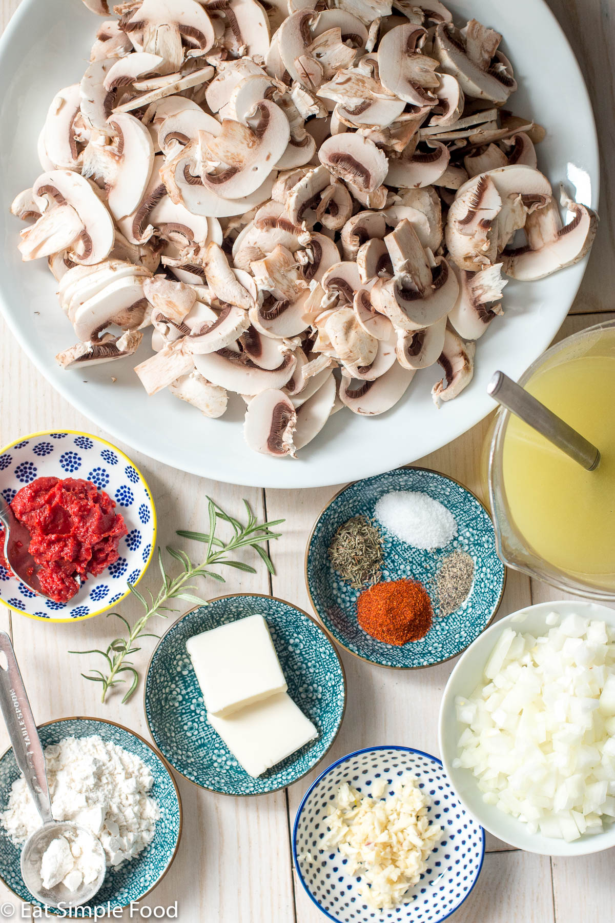 Ingredients on a white table: plate of sliced mushrooms, tomato paste, rosemary sprig, 2 tabs of butter, container of flour with 1 tablespoon in it, diced onion bowl, minced garlic bowl, small bowl of mixed spices, chicken broth with spoon.