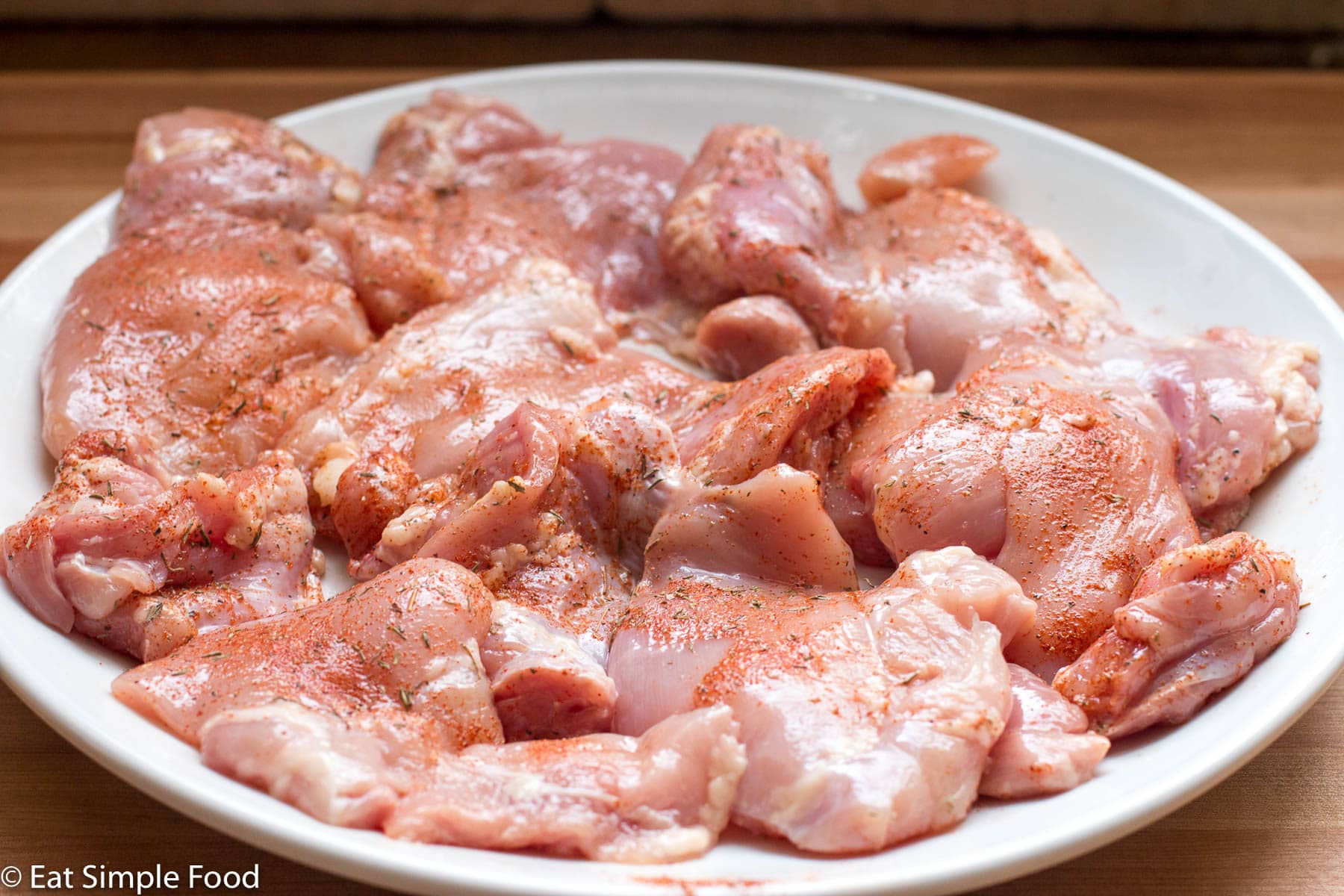 White plate of raw boneless and skinless chicken thighs with spices.