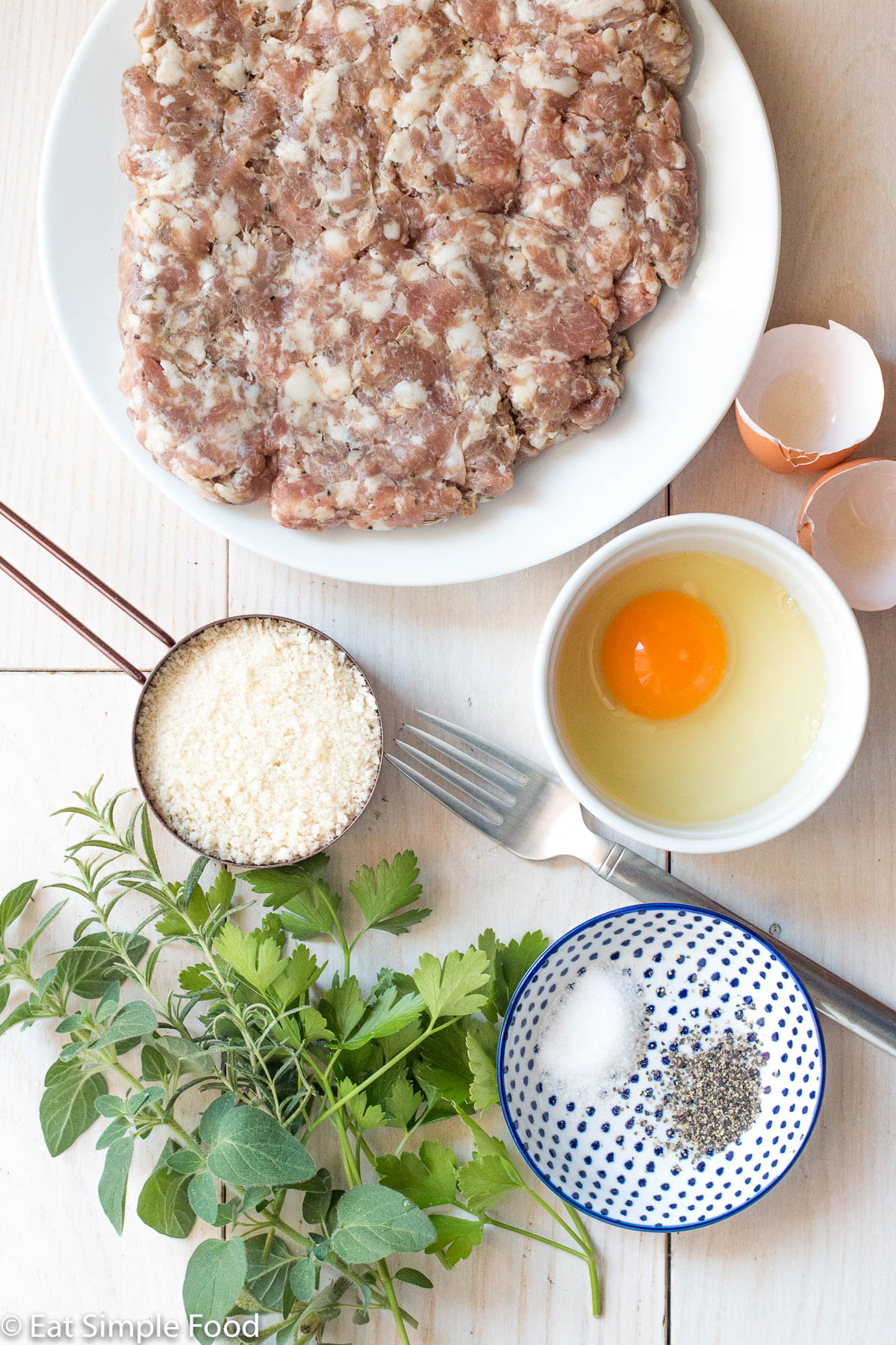 Ingredients on a white table: white plate of ground sausage, one cracked egg in a white bowl, ½ cup panko bread crumbs, container of salt and pepper, and fresh herbs (oregano, rosemary, parsley).