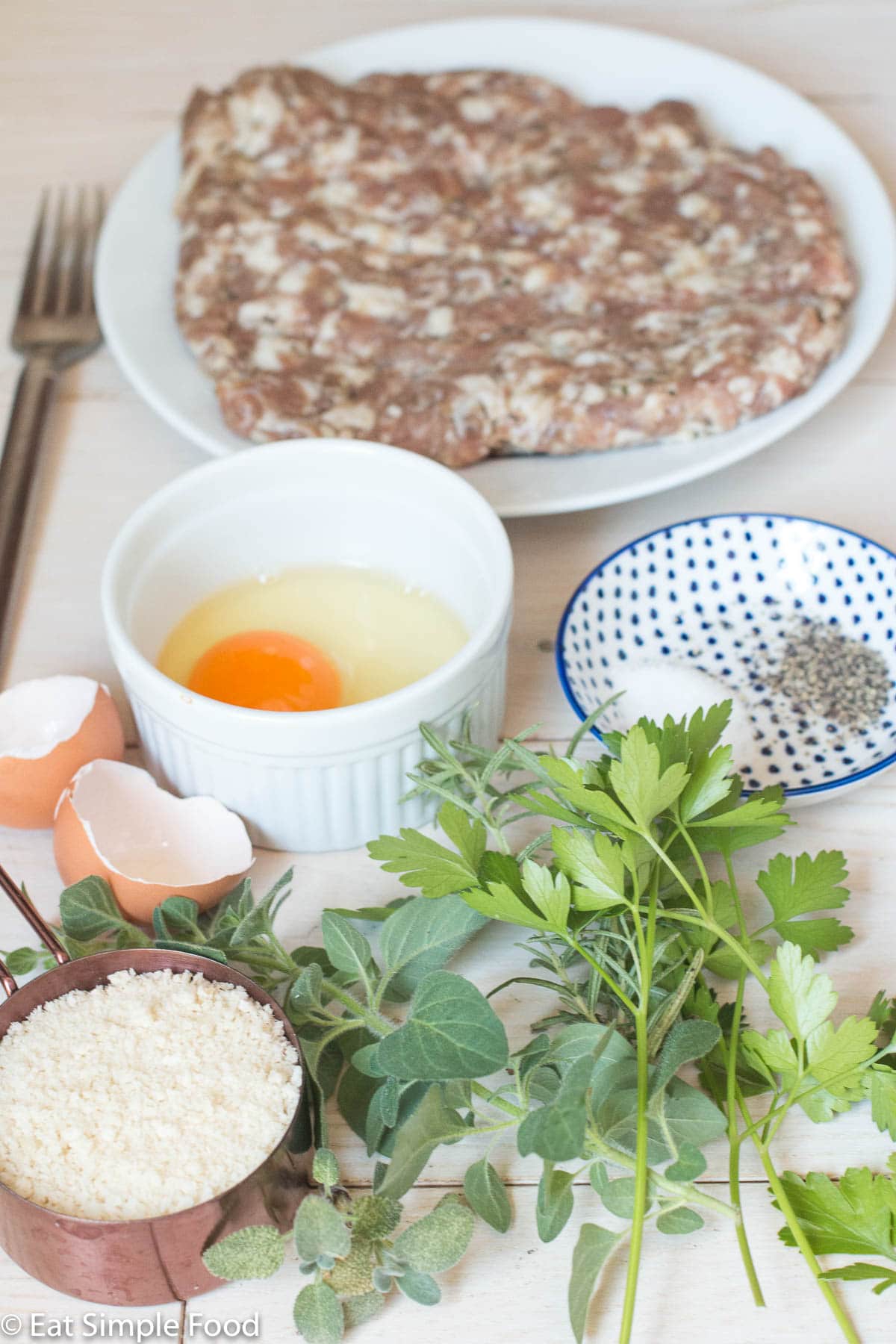 Ingredients on a white table: white plate of ground sausage, one cracked egg in a white bowl, ½ cup panko bread crumbs, container of salt and pepper, and fresh herbs (oregano, rosemary, parsley). Side view.