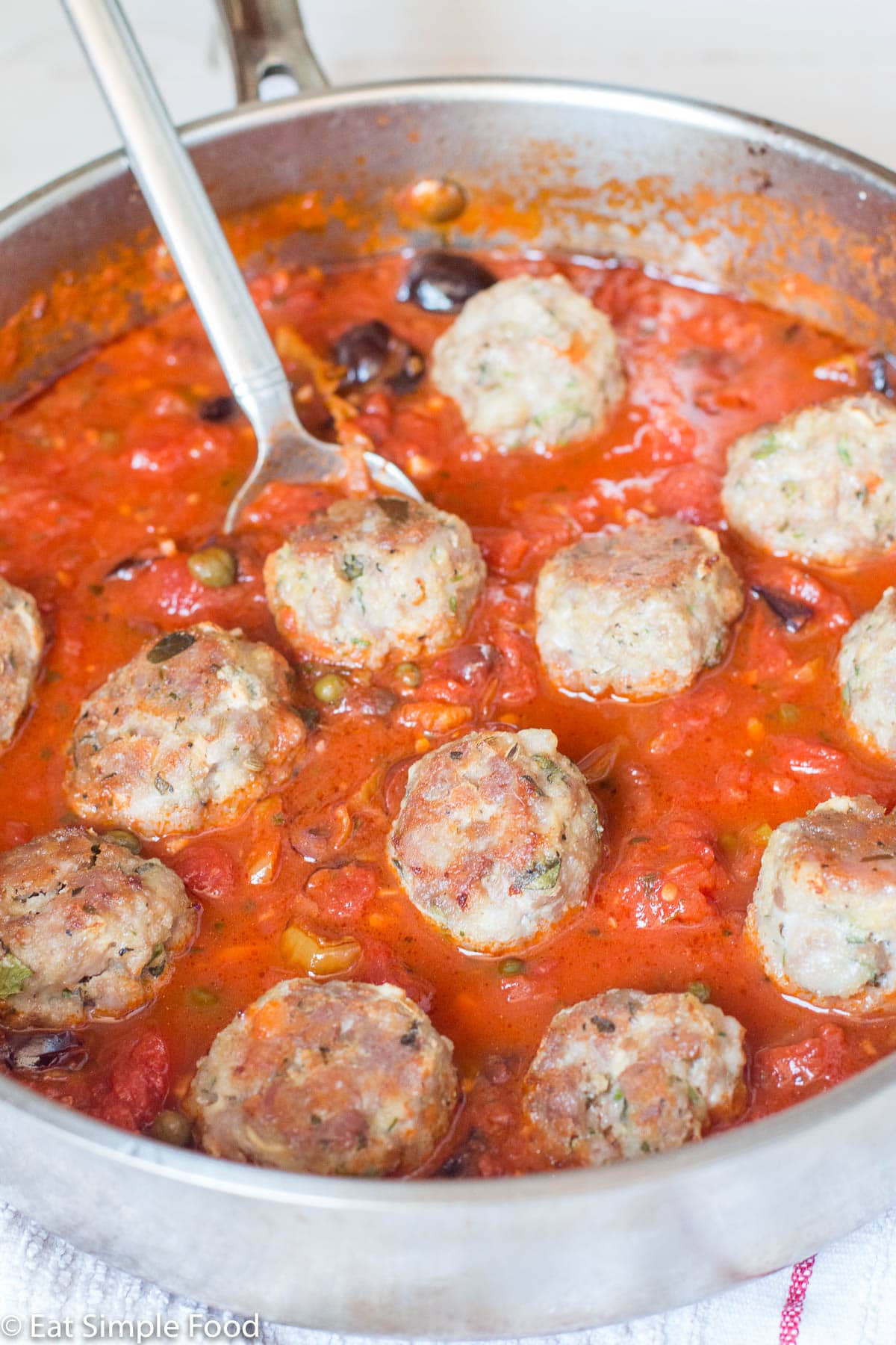 Large stainless steel pan with a silver spoon. Pan is full of browned sausage meatballs in a tomato sauce with olives and capers. Close up.