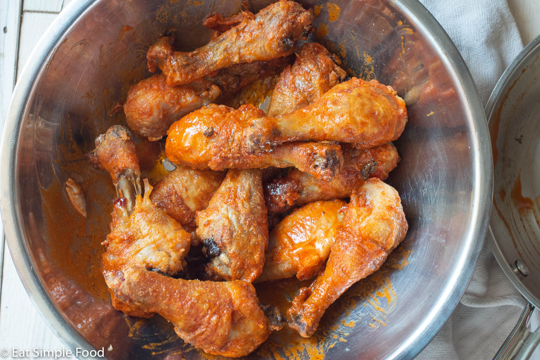 12 baked chicken drumsticks with a red/orange hot sauce coating tossed in a stainless steel bowl. Top view.