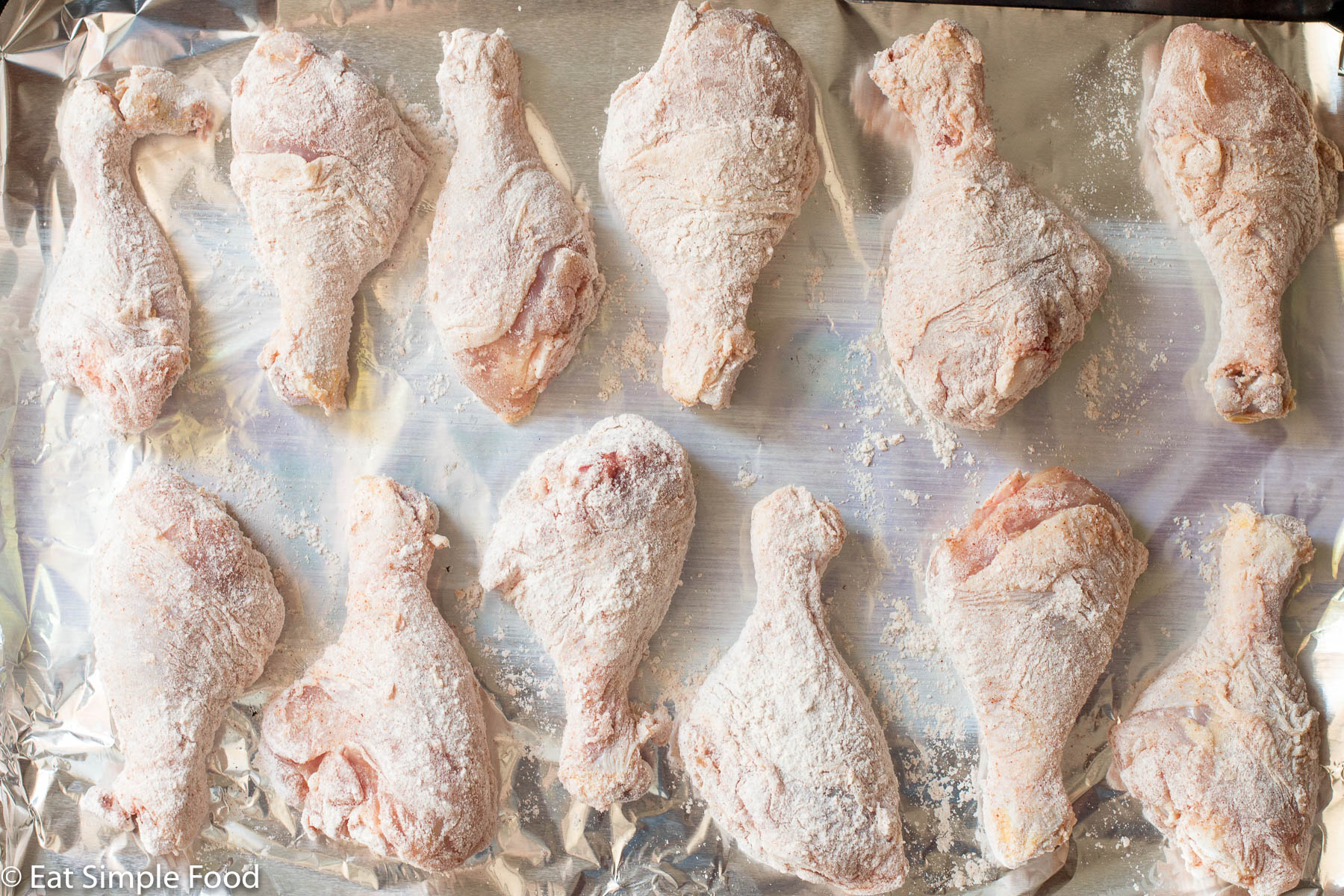 12 Raw Chicken drumsticks on an aluminum covered baking sheet. Top view.