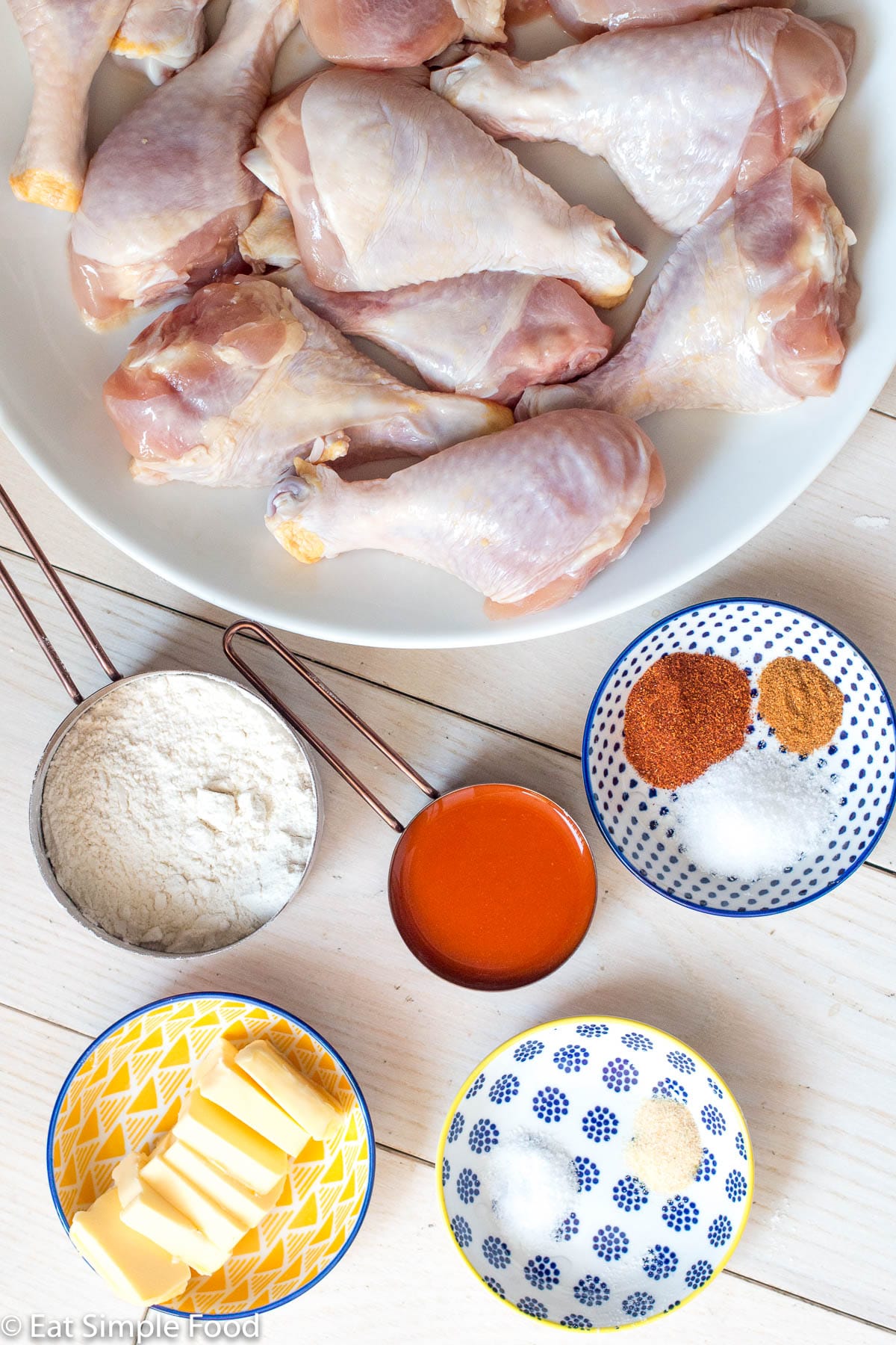 Raw chicken drumsticks on a white plate with hot sauce, butter, flour, and paprika in containers around it. Top view.