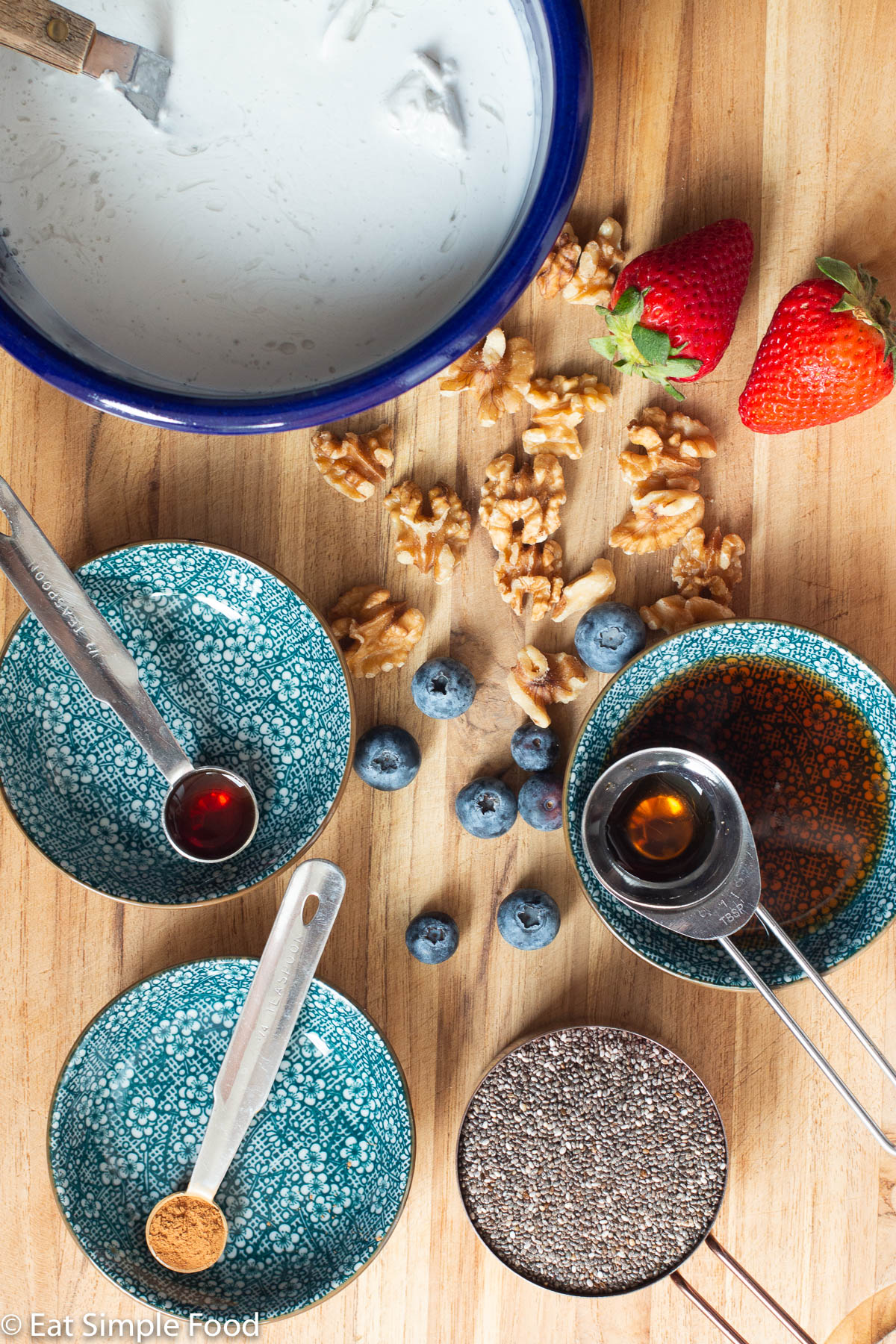 Top view of ingredients in small blue bowls on a wood cutting board: walnuts, blueberries, 2 strawberries, coconut milk, chia seeds, cinnamon, maple syrup, vanilla extract.