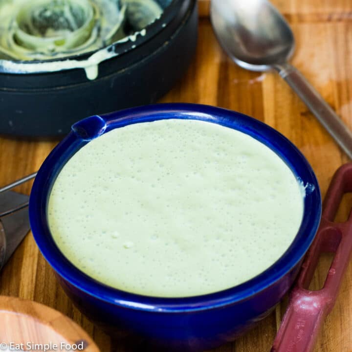 Blue bowl of green cilantro sour cream sauce on a wood cutting board with a red spatula on the side. Blender top in the background with green sauce all over it. Close up.