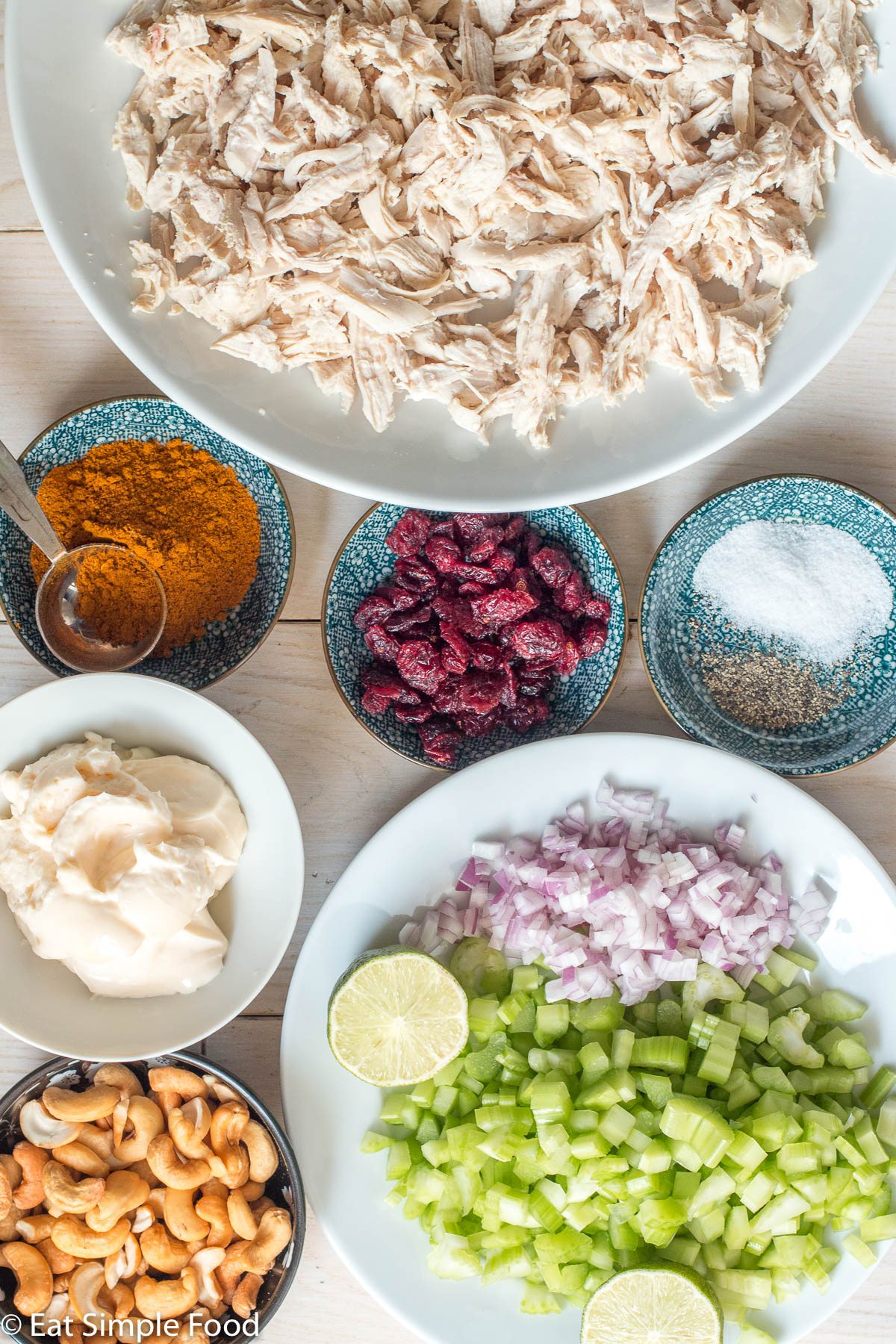Top view of ingredients: white plate of shredded chicken, bowl of cranberries, bowl of curry powder, bowl of salt and pepper, bowl of mayonnaise, bowl of cashews, plate of diced celery and red onions with a lime cut in half.