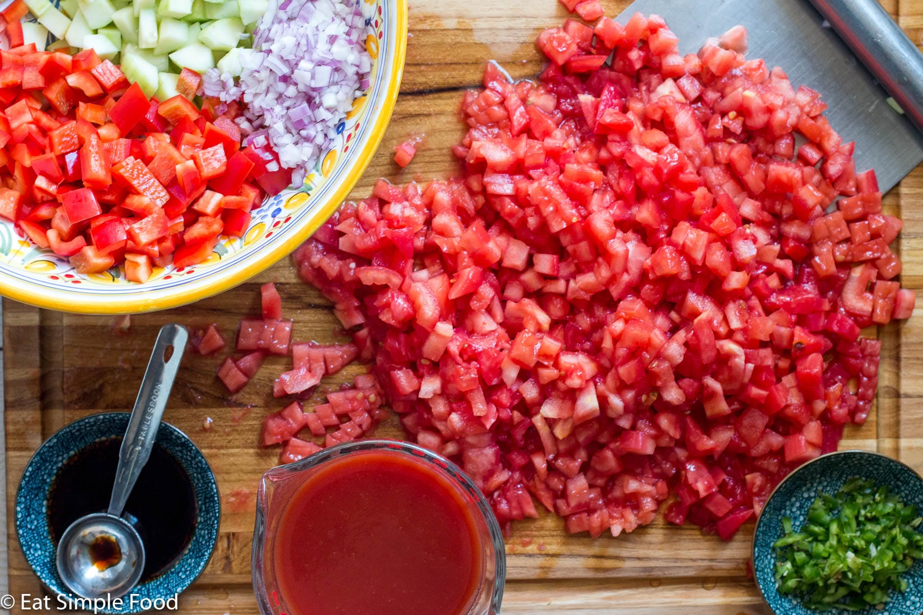 Chopped fresh tomatoes on a wood cutting board with a colorful bowl of chopped red peppers, red onions, cucumbers, and jalapenos on the side.