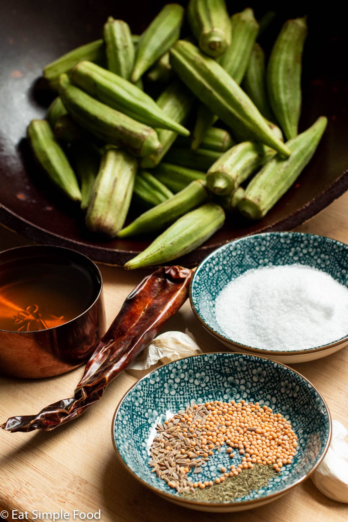 Ingredients on a wood cutting board: whole okra in brown bowl, dried red chile, garlic clove, olive oil container, salt, and small blue spice container with mustard seeds, dill seeds, and dried dill.
