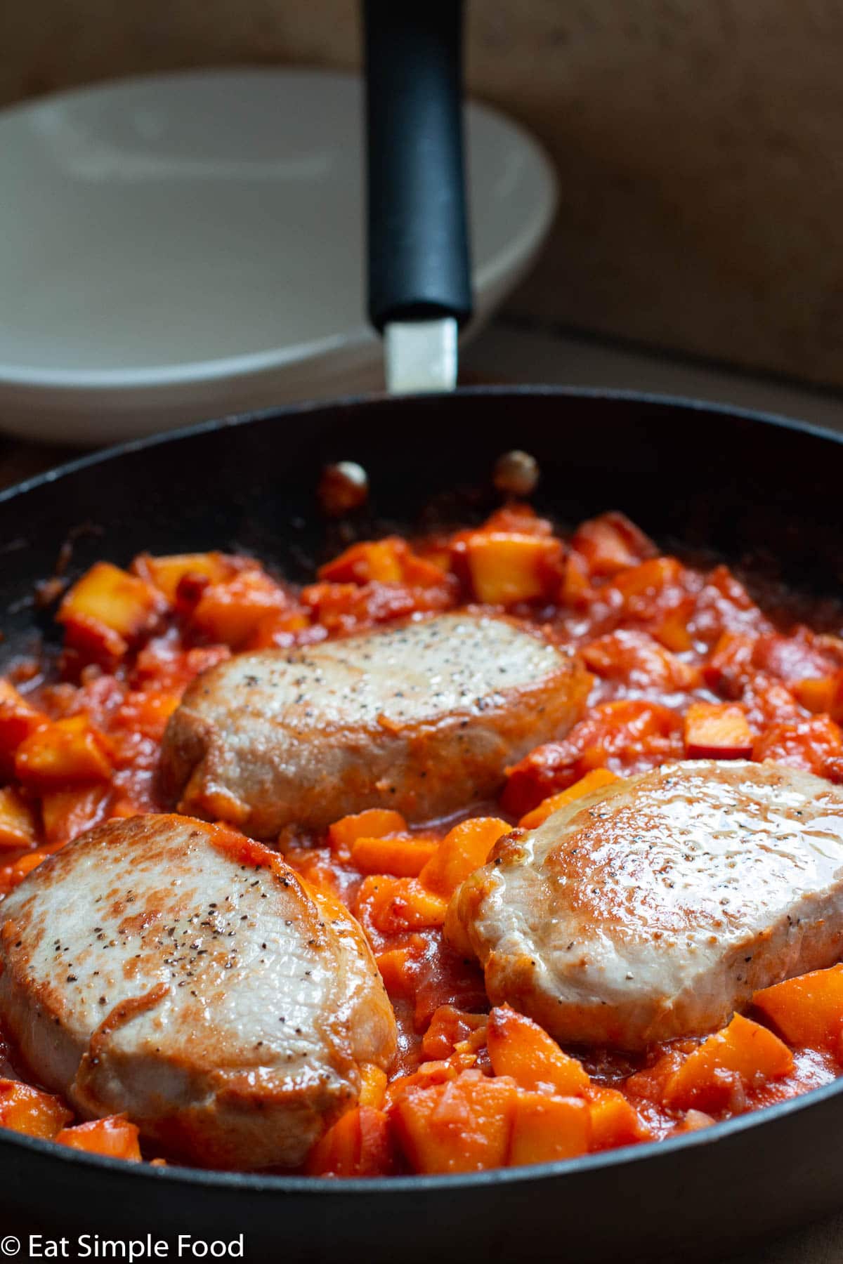 Side view of 3 boneless pork chops in a peach tomato sauce in a black skillet.