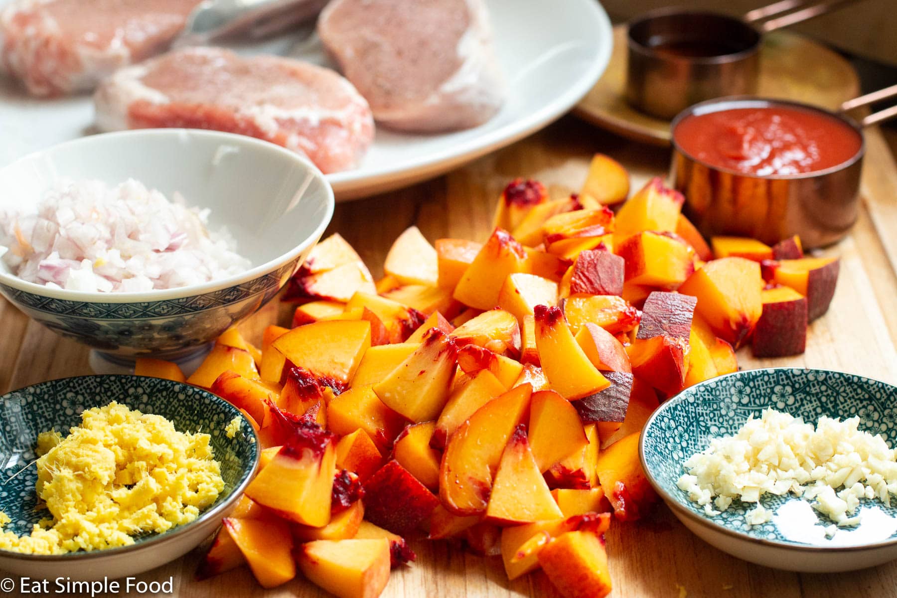 Diced fresh peaches on a wood cutting board with other ingredients: diced garlic, minced ginger, diced shallots, tomato sauce, and 3 raw pork chops in background on a white plate.