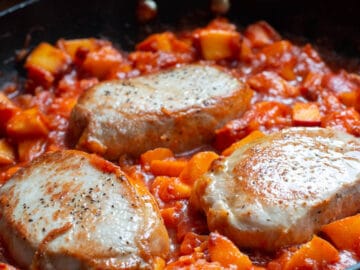Side view of three boneless pork chops in a peach tomato sauce in a black skillet. Close up.