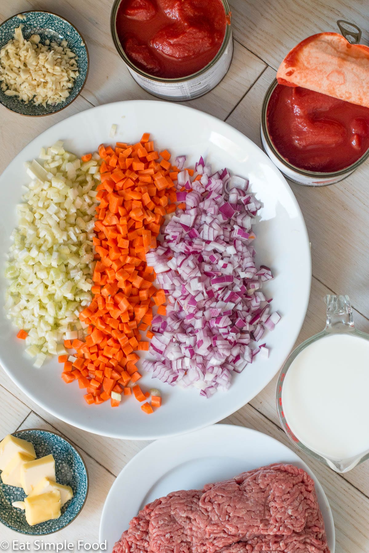 Raw ingredients on white plates and colorful bowls: chopped red onions, chopped carrots, chopped celery, raw ground beef, butter tabs, minced garlic, 2 cans of tomatoes, and glass of milk.