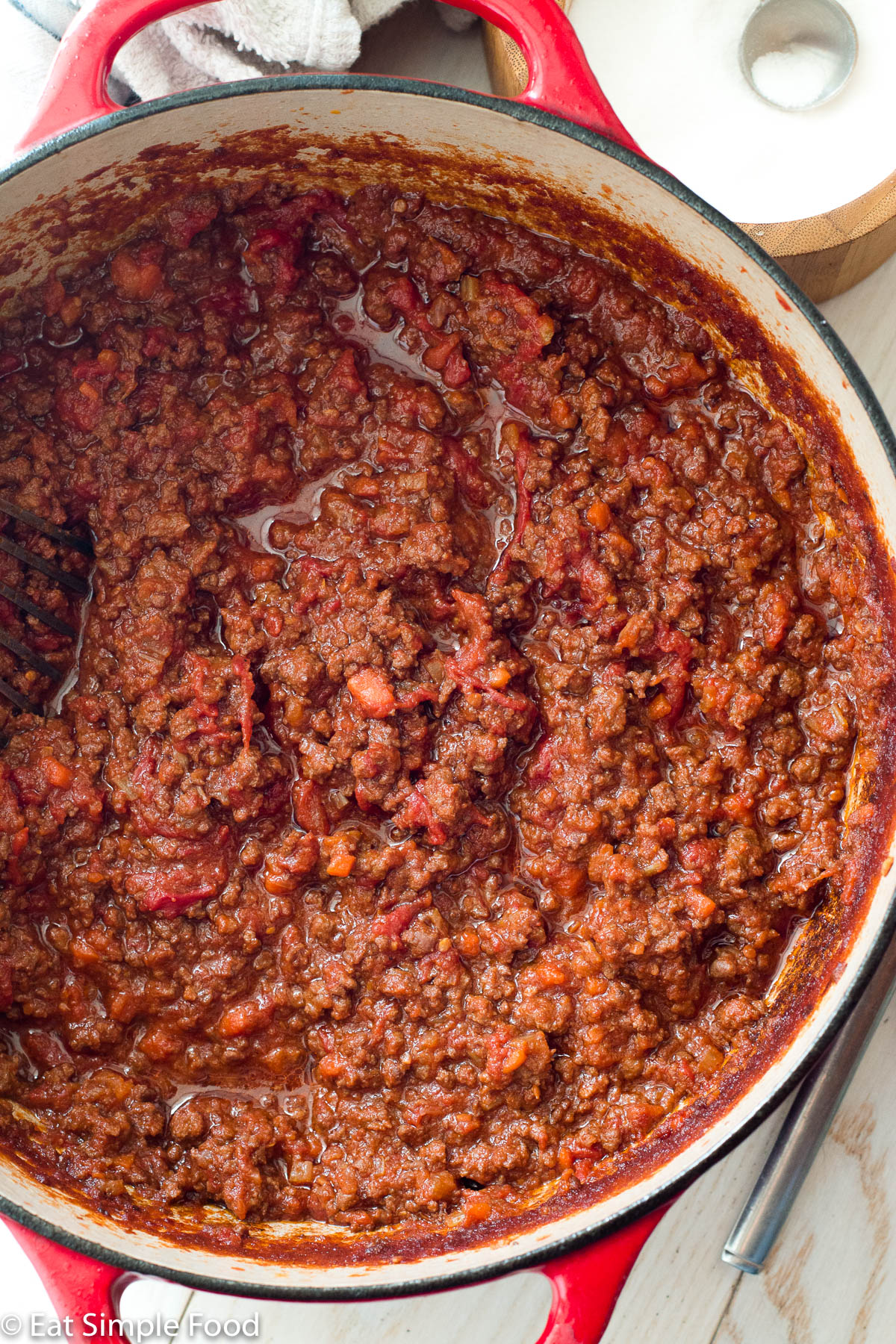 Red cast iron pot filled with meat and tomato bolognese sauce. Top view. Salt bowl on side.