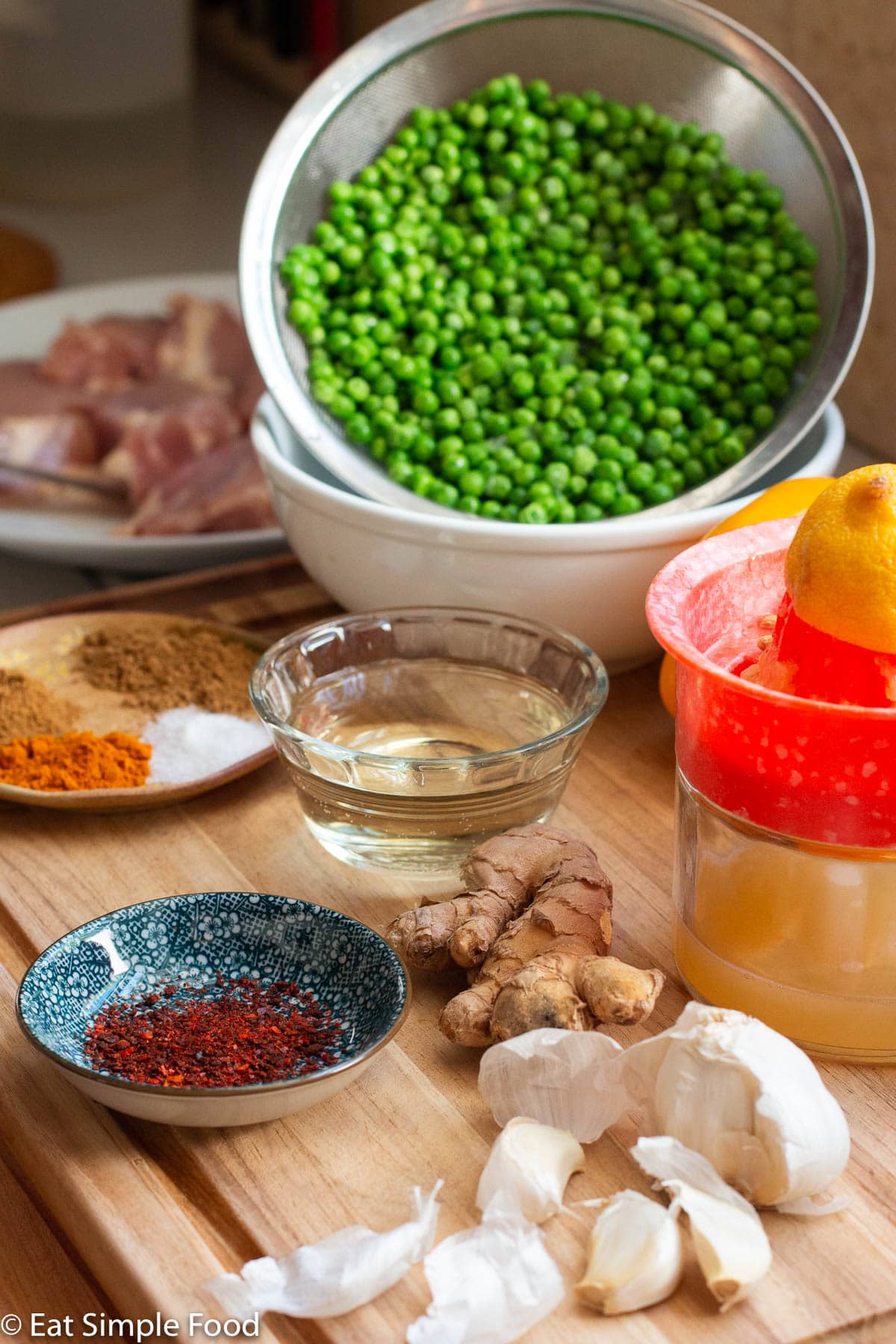 Raw ingredients on a wood cutting board: garlic cloves, lemon juice and juicer with lemon on it, ginger knob, bowl of white wine, bowl of chili pepper flakes, small plate of spices, strainer of vibrant green peas.