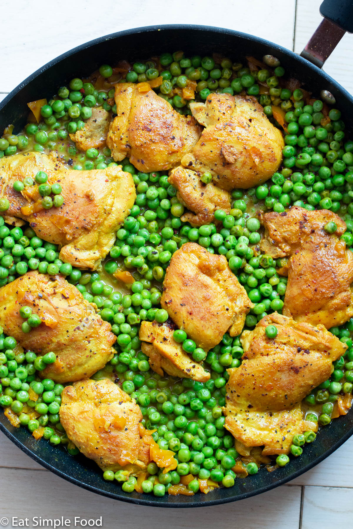 Top view of browned boneless skinless chicken thighs on a bed of vibrant green peas in a black skillet.