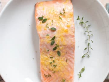 One piece of cooked salmon on a white plate. Salmon is topped with a lemon zest butter and fresh thyme. On a white plate with colorful napkin on the side. Close up.