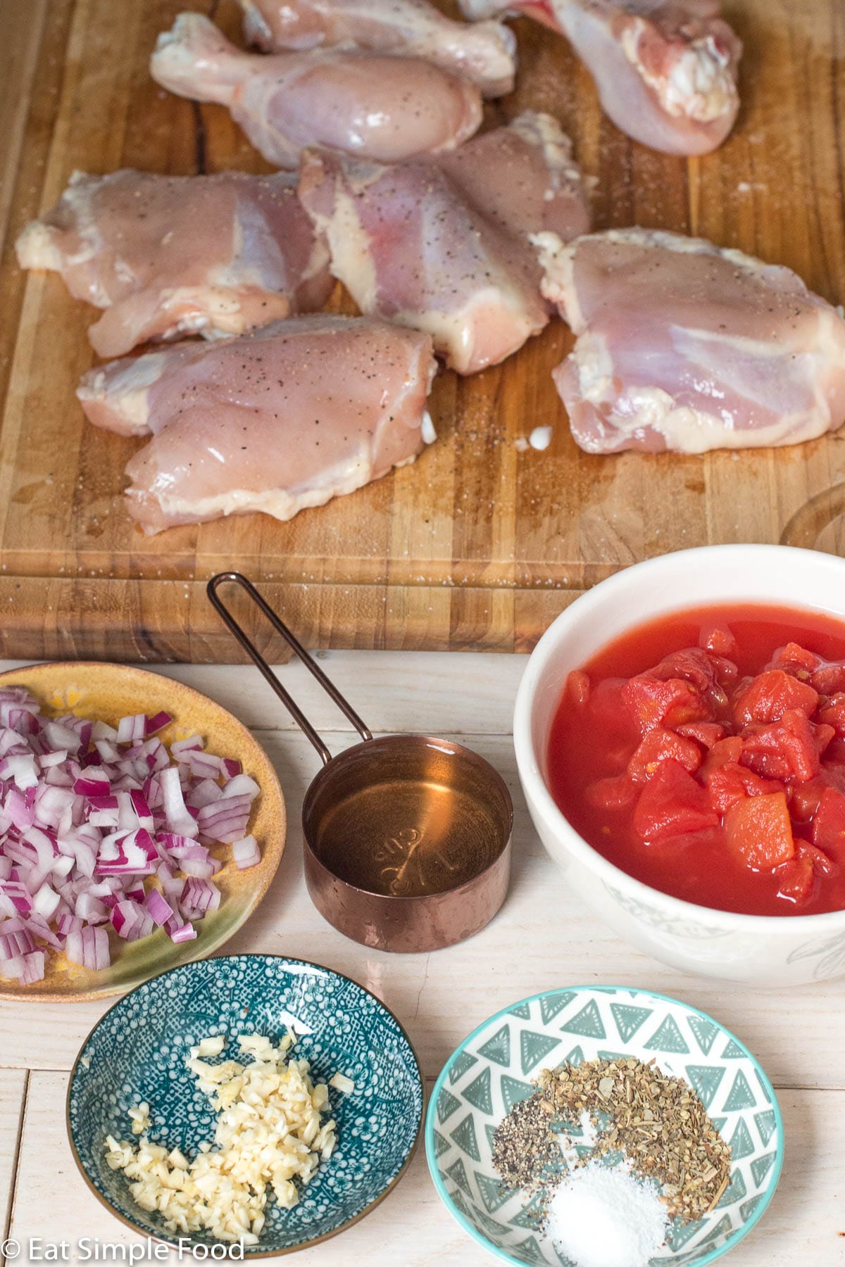 Raw ingredients: diced red onions, minced garlic, bowl of diced canned tomatoes, ⅓ cup white wine, raw chicken thighs and drums on a wood cutting board. Side view.