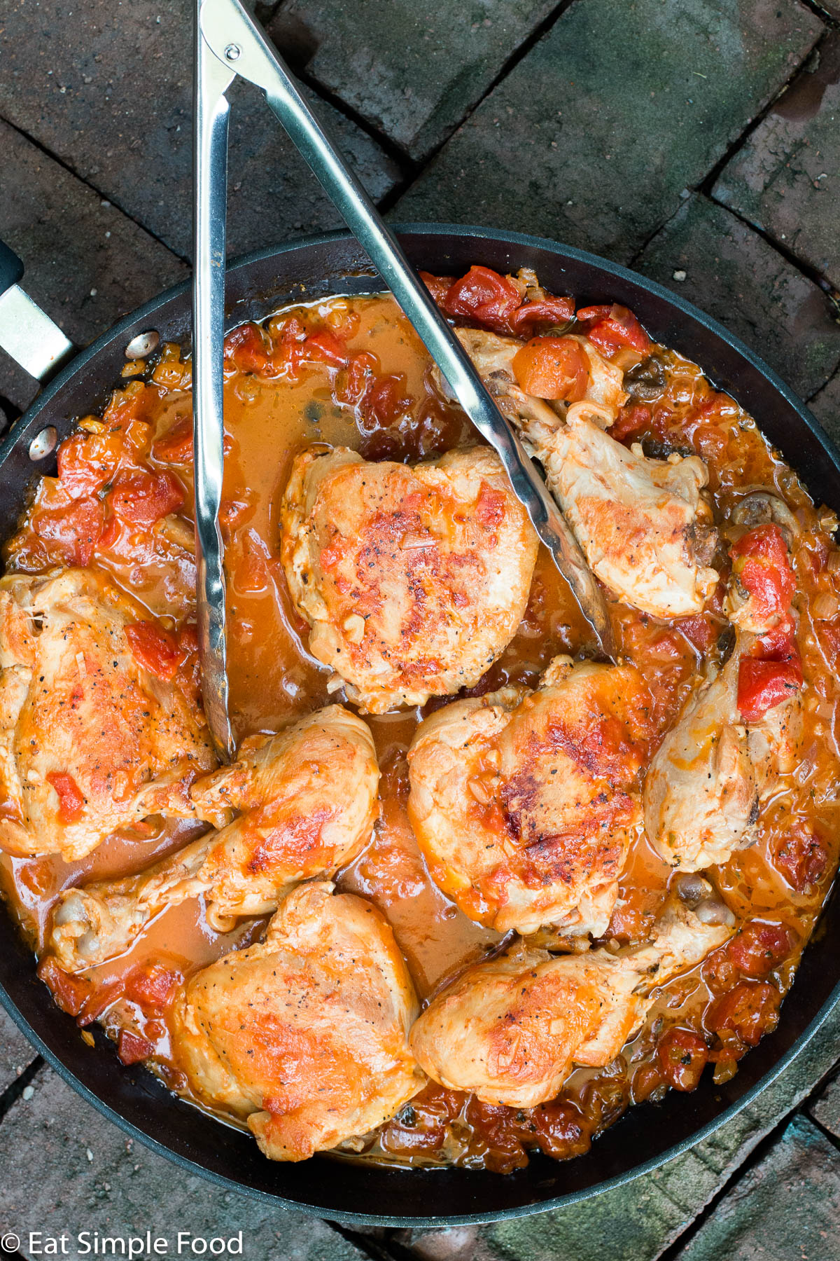 Top down view of browned chicken in a tomato and white wine sauce in a large black pan with tongs hanging out the side.