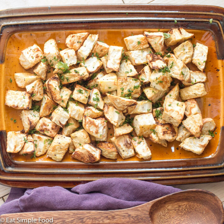 Crispy roasted chunks of celery root on a brown platter with a wood spoon and a green garnish. Top down view.