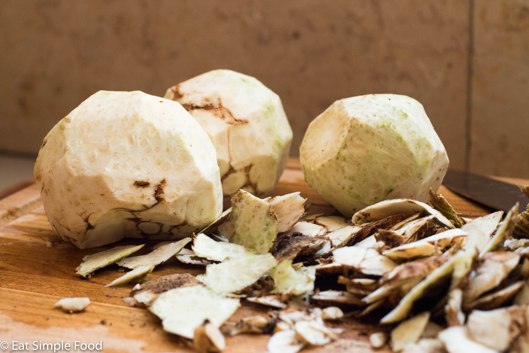 3 peeled celery root (celeriac) bulbs on a wooden cutting board with the peels on the side.