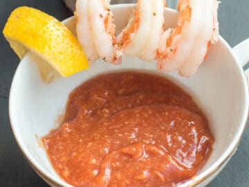 White tea chup with 3 poached shrimp and a lemon wedge on the edge with cocktail sauce in the cup. Close up.