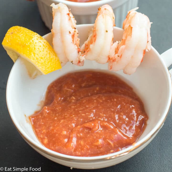 White tea chup with 3 poached shrimp and a lemon wedge on the edge with cocktail sauce in the cup. Close up.