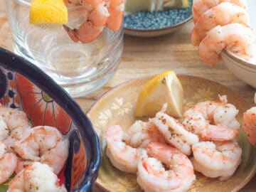 Five poached pink shrimp on a little tan plate with a wedged lemon. Poached shrimp in a blue bowl on the side and also layed over some cups in the background. Lemon wedges in the background.