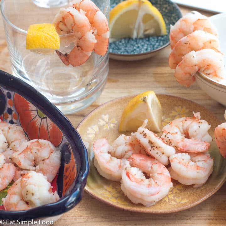 Five poached pink shrimp on a little tan plate with a wedged lemon. Poached shrimp in a blue bowl on the side and also layed over some cups in the background. Lemon wedges in the background.