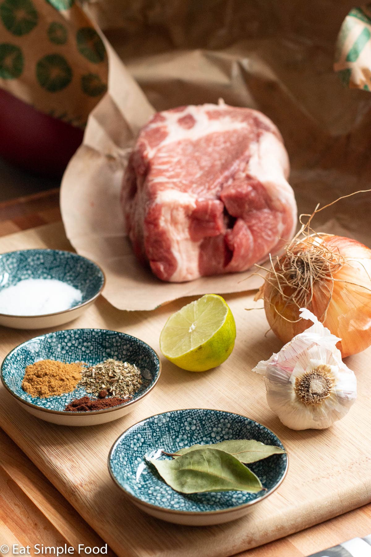 Raw ingredients on a wood cutting board: chunk of boneless pork shoulder, lime half, 2 bay leaves, 4 colorful spices, garlic knob, and whole onion.