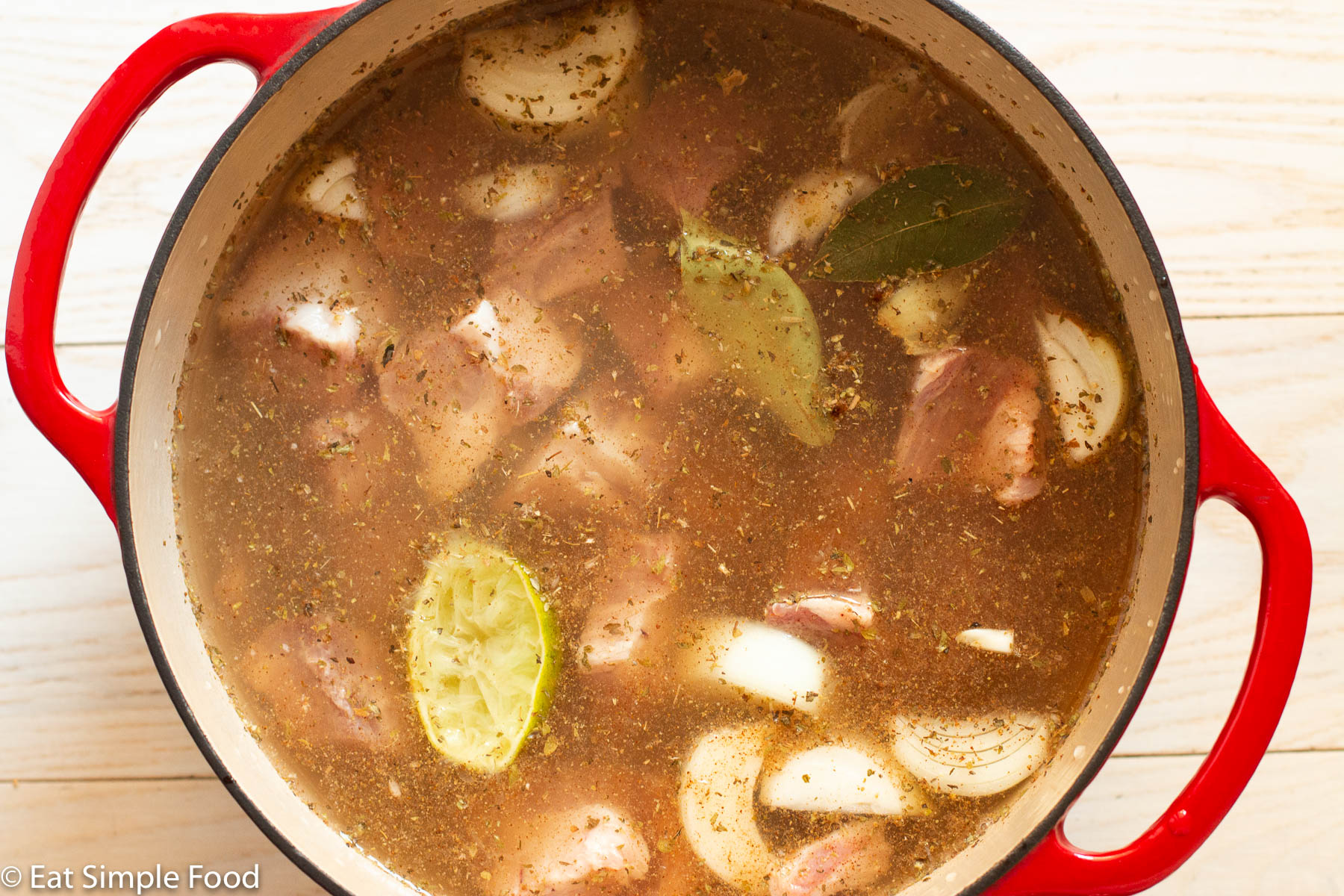 Red stockpot of uncooked chunked pork, lime halves, broth, herbs, onion chunks, and two bay leaves.