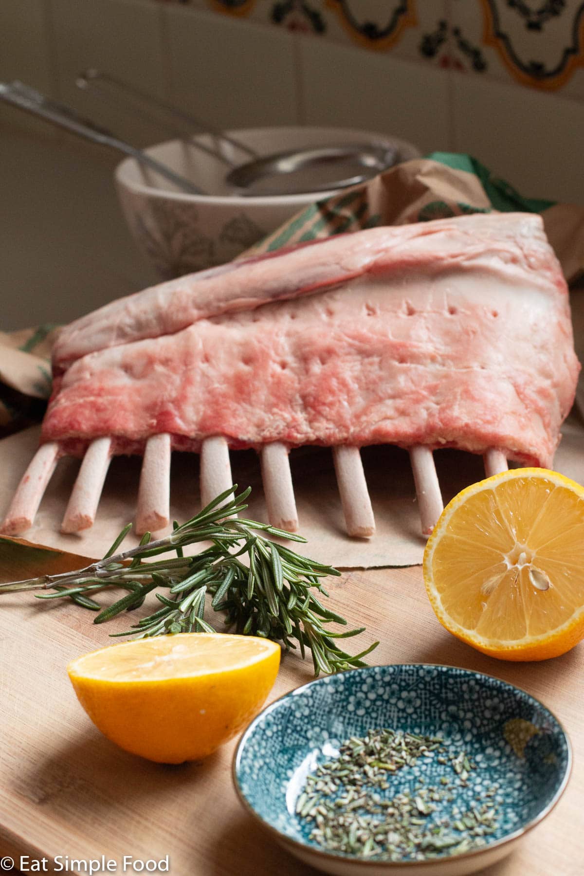 Raw ingredients: 8 bone rack of lamb, 2 lemon halves, diced rosemary in a container and a large rosemary sprig on a wood cutting board.