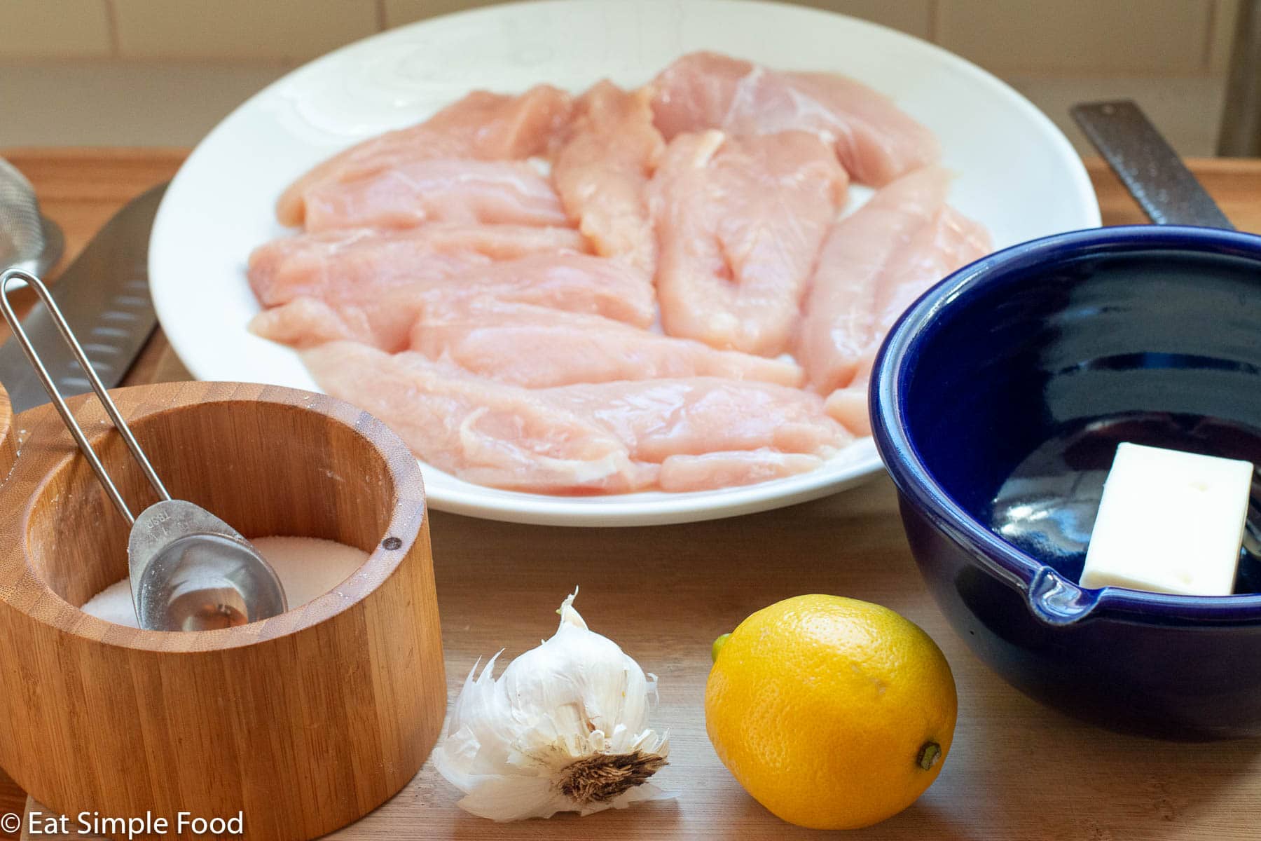 Raw chicken on a white plate and ingredients in front: wood bowl of salt, whole lemon, whole garlic, blue bowl of stick of butter.