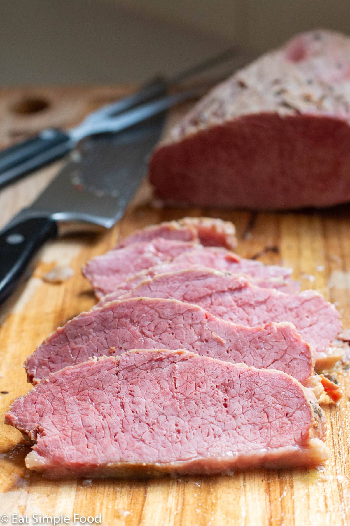 Side view of slices of think pink corned beef on a wood cutting board with a knife in the background.