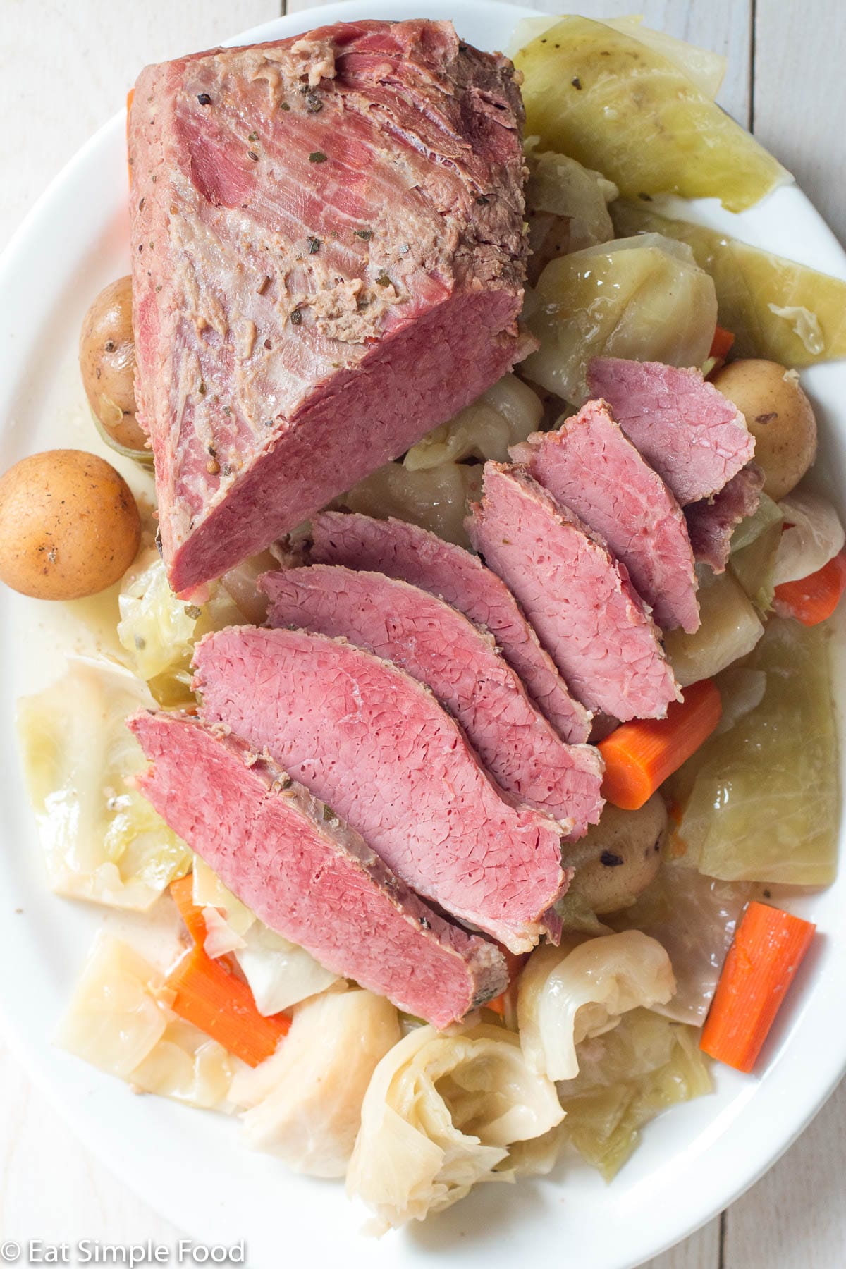 Top down view of a chunk of corned beef with layered pink thin slices on a bed of cooked cabbage, carrots, and baby potaotes.
