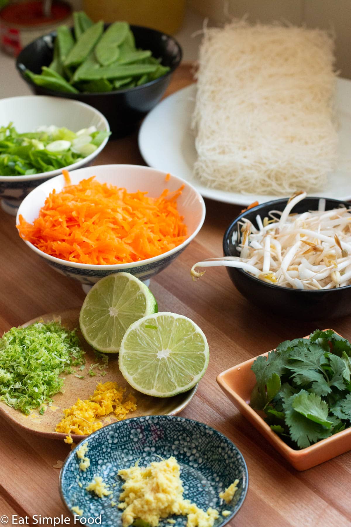 Raw cut up and sliced veggies on a wood cutting board in bowls: shredded carrots, lime zest, lemon zest, minced ginger, cilantro leaves, mung beans, uncooked rice noodles, sugar snap peas, and sliced green onions.