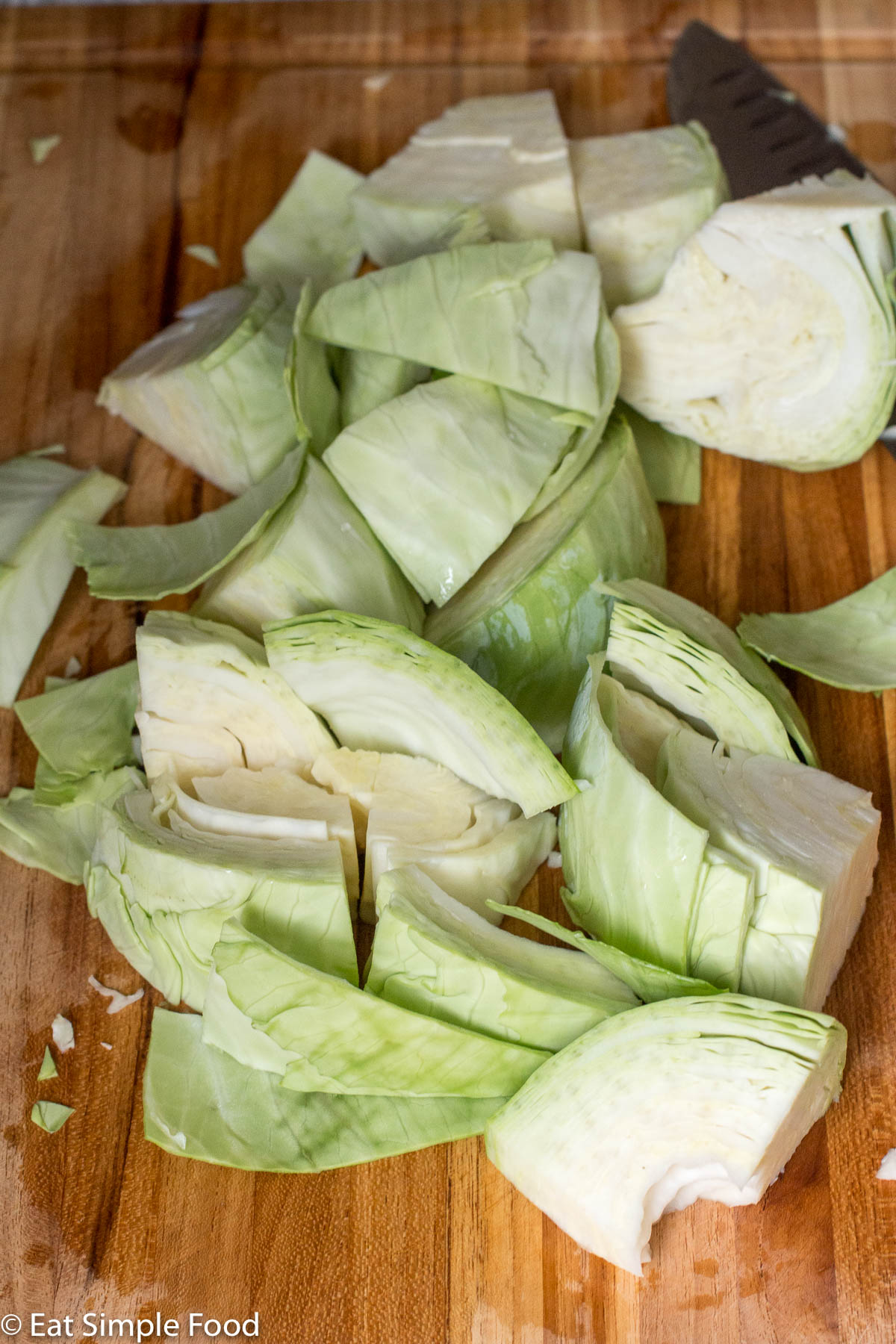 Chunks of raw cut up cabbage on a wood cutting board with a chefs knife in the background.